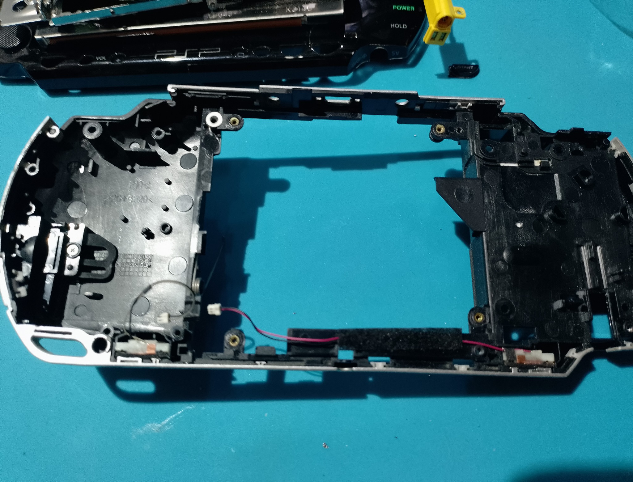 PSP-1004 замена корпуса - Старый чёрный нижний корпус снят с консоли. Внутри остаются провод динамика, посадочные места и следы прошлой эксплуатации.